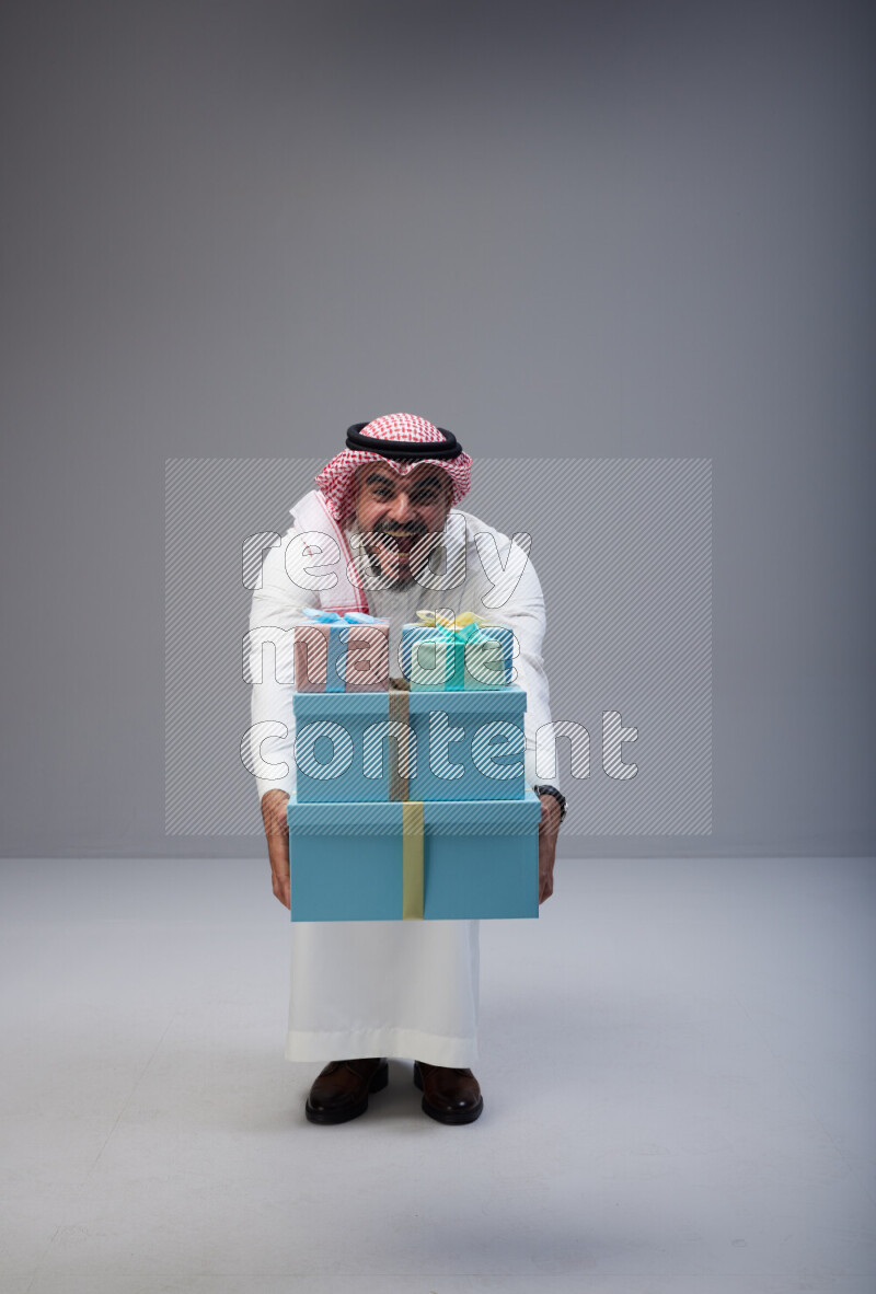 Saudi man Wearing Thob and red Shomag standing holding gift box on Gray background