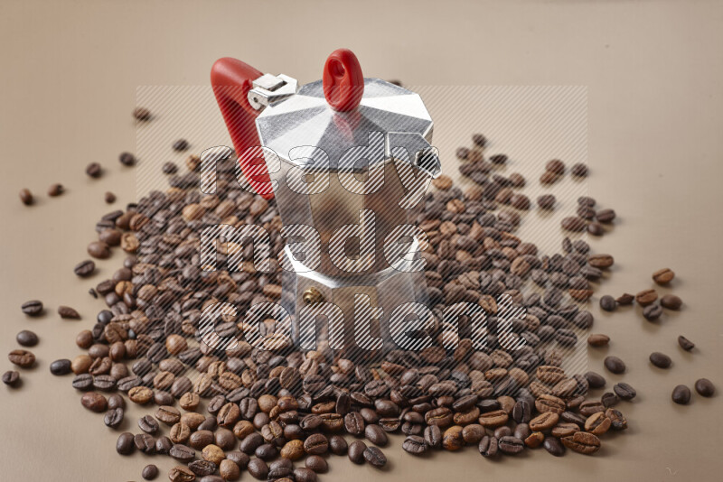 A moka pot with red handle surrounded by roasted coffee beans on beige background
