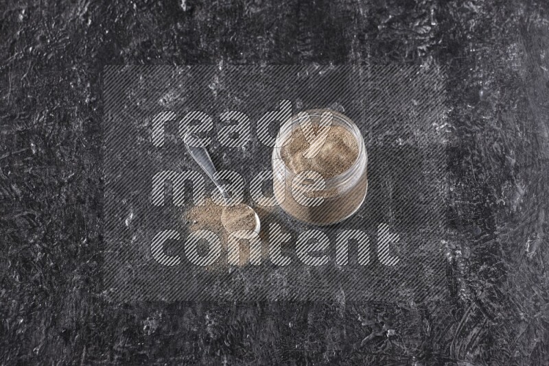 A glass jar full of black pepper powder and a metal spoon full of powder on a textured black flooring