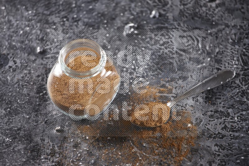 Herbal glass jar full of cinnamon powder and a metal spoon full of powder on textured black background