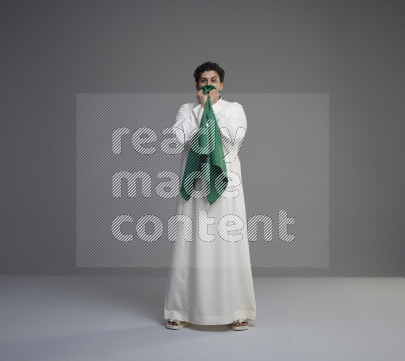 A Saudi man standing wearing thob kissing Saudi flag on gray background
