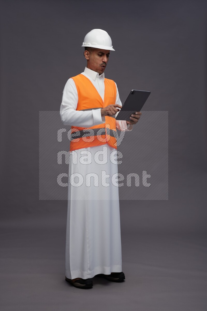 Saudi man wearing thob with engineer vest standing working on tablet on gray background
