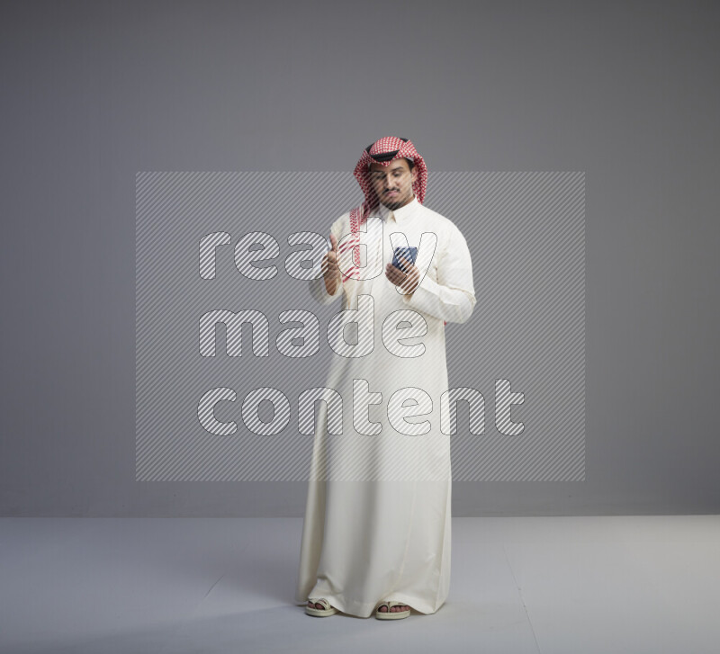 A  Saudi man standing wearing thob and red shomag texting on phone on gray background