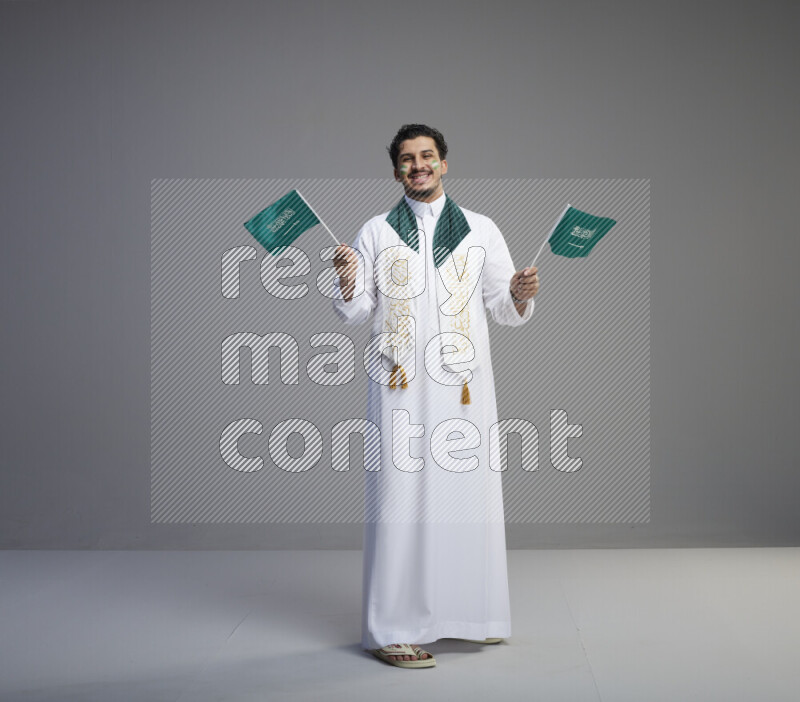 A Saudi man standing wearing thob and saudi flag scarf with face painting holding small Saudi flag on gray background