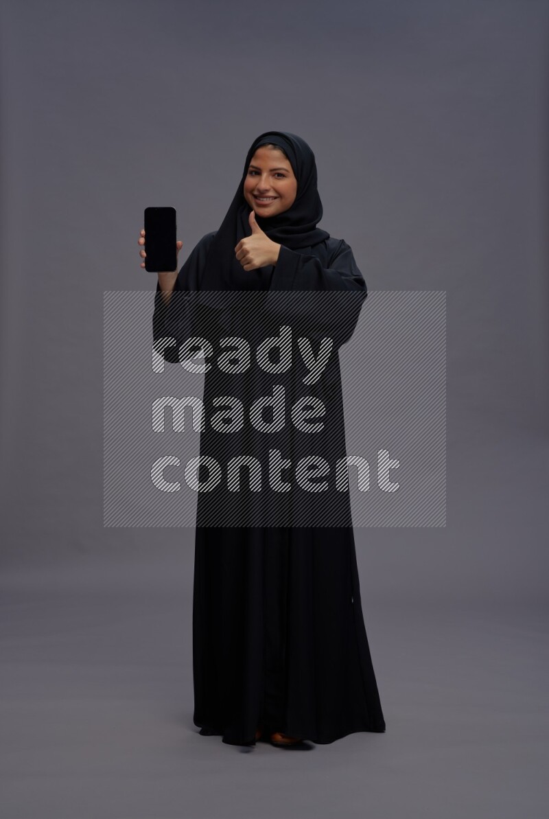 Saudi woman wearing Abaya standing showing phone to camera on gray background