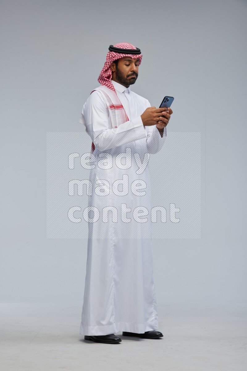 Saudi man Wearing Thob and shomag standing texting on phone on Gray background