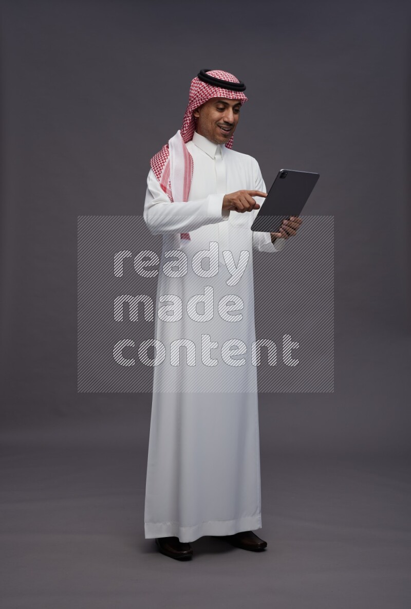 Saudi man wearing thob and shomag standing working on tablet on gray background
