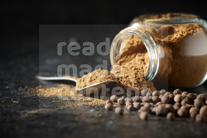 A flipped glass spice jar and metal spoon full of allspice powder and powder spilled out of it with whole balls on a textured black flooring
