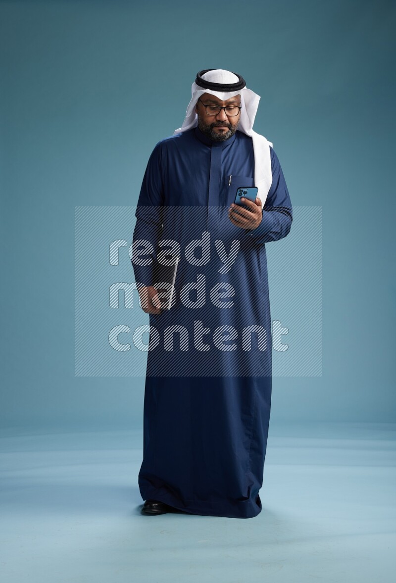 Saudi Man with shimag Standing texting on phone on blue background