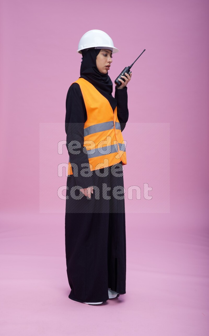Saudi woman wearing Abaya with engineer vest and helmet standing holding walkie-talkie on pink background