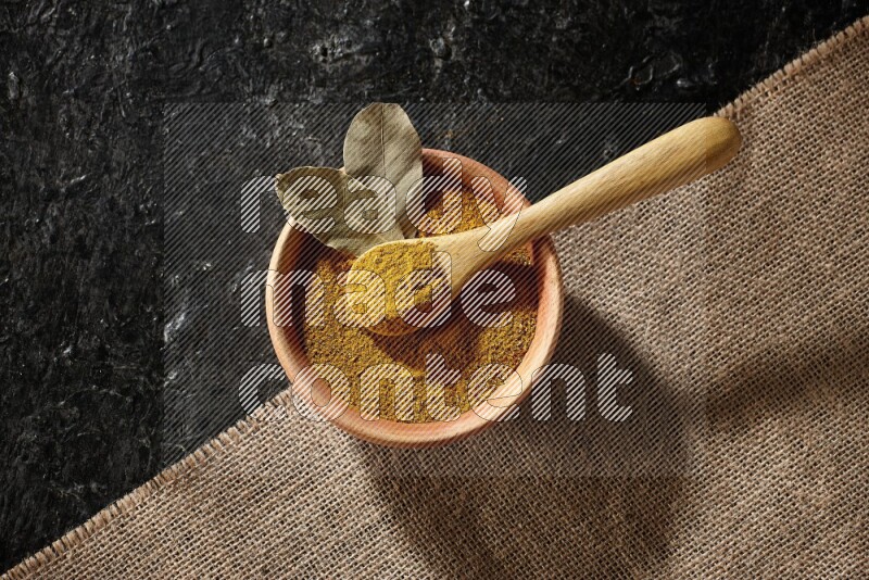 A wooden bowl and a wooden spoon full of turmeric powder on burlap fabric on textured black flooring