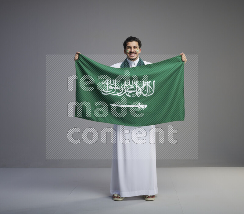 A Saudi man standing wearing thob with face painting wrapping Saudi flag scarf and holding big Saudi flag on gray background
