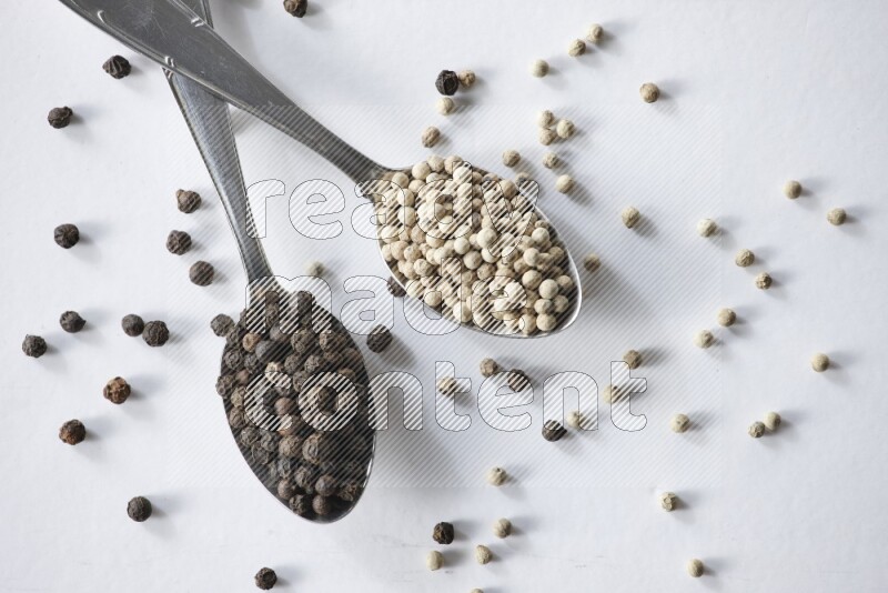 2 metal spoons full of black and white pepper beads with spreaded beads on white flooring