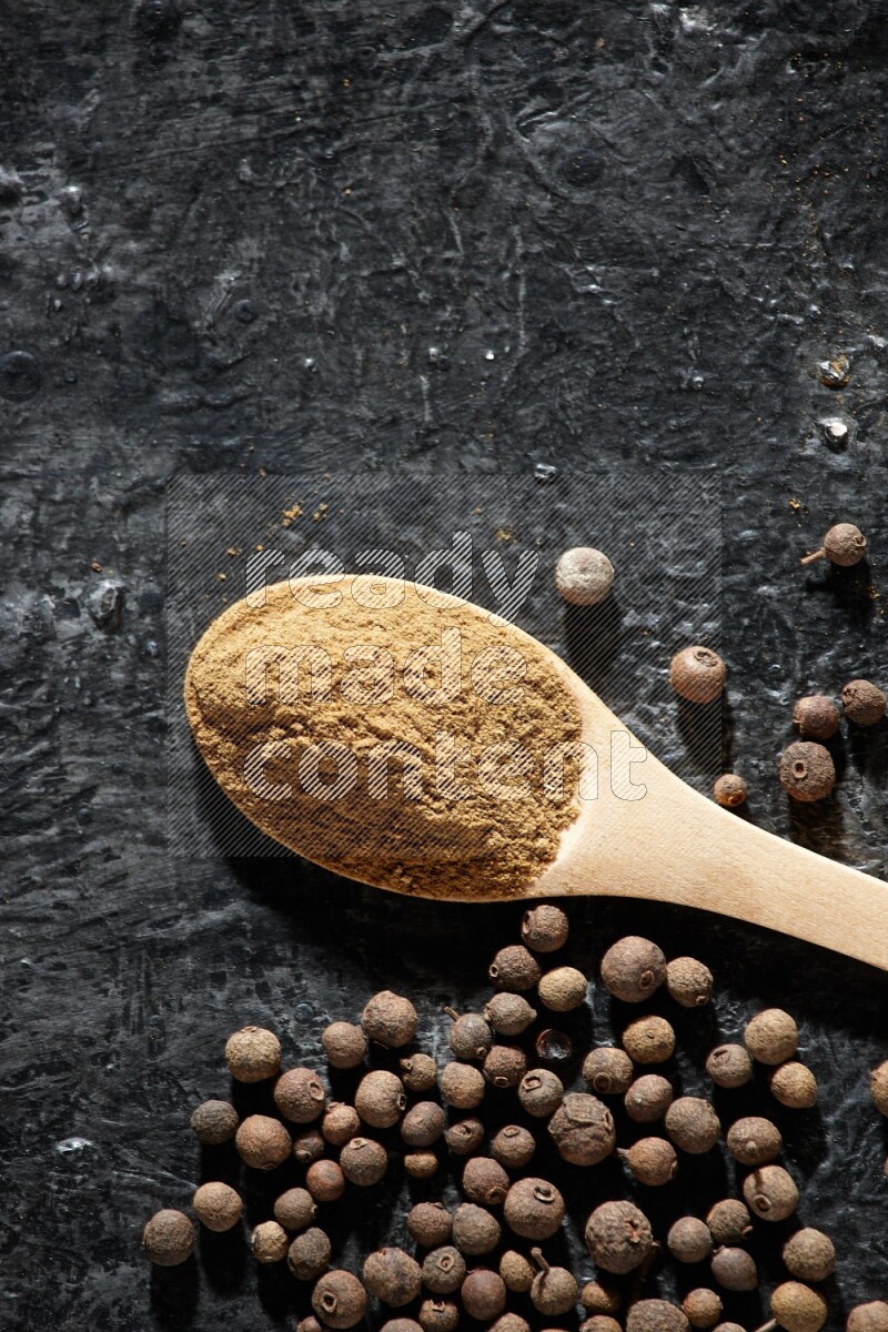 Wooden spoons full of all spice powder and allspice whole balls beside it on a textured black flooring