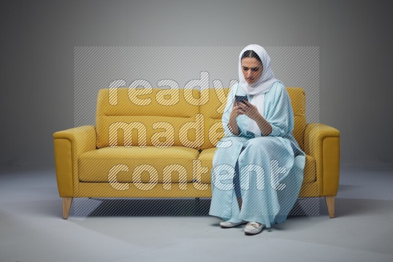 A Saudi woman wearing a light blue Abaya and white head scarf sitting on a yellow sofa and texting on her phone on a grey background