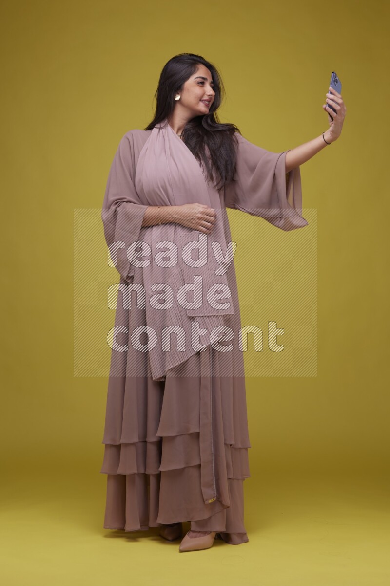 A woman Taking a Selfie on a Yellow Background wearing Brown Abaya