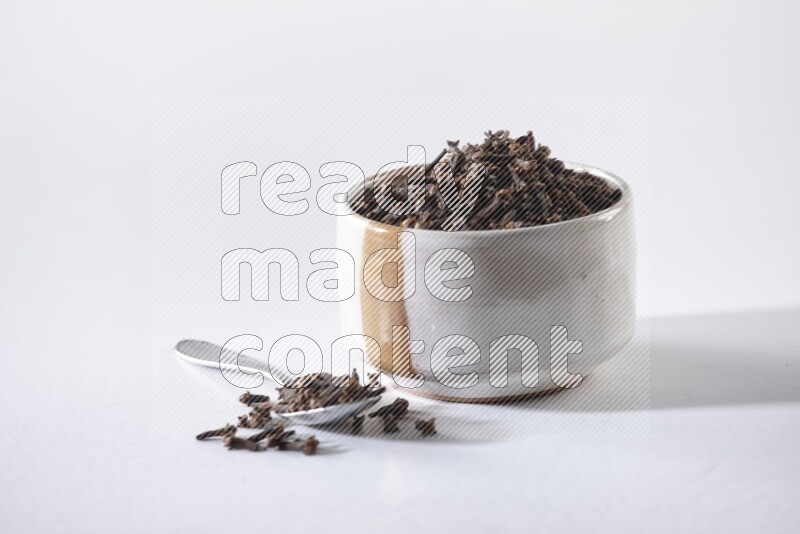 A beige ceramic bowl full of cloves and a metal spoon next to it on a white flooring