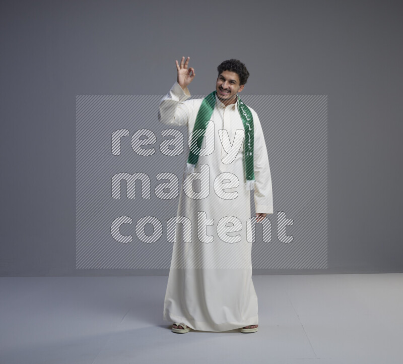 A Saudi man standing wearing thob and Saudi flag scarf on gray background