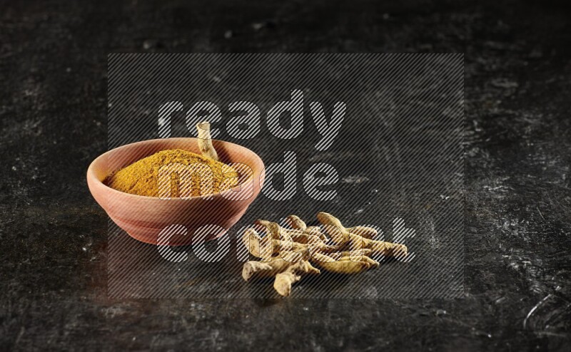 A wooden bowl full of turmeric powder with dried turmeric whole fingers on textured black flooring
