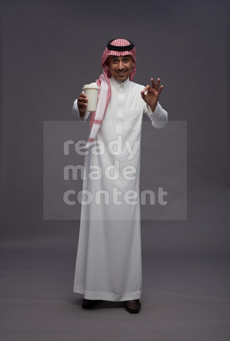 Saudi man wearing thob and shomag standing holding paper cup on gray background