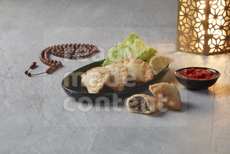 Three fried sambosas in an oval shaped black plate and a red sauce in a black round ramekin with a brown misbaha and a golden lantern on the side on a gray background