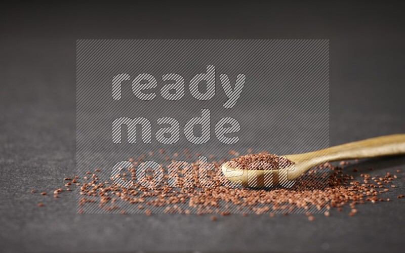 A wooden spoon full of garden cress seeds and seeds spread beside it on a black flooring