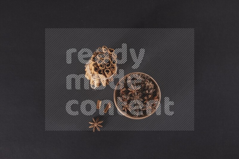 A stacked and bounded cinnamon sticks and a wooden bowl full of star anise on a black background