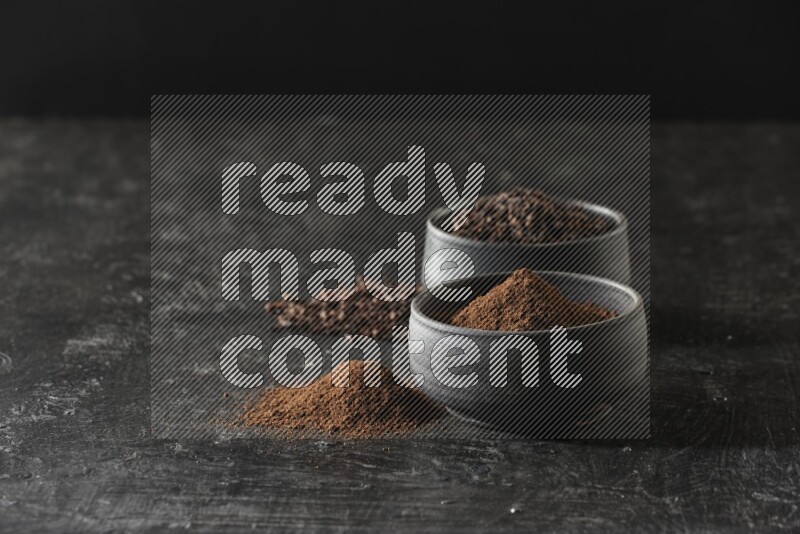 2 Black pottery bowls full of cloves and the other full of cloves powder on textured black flooring