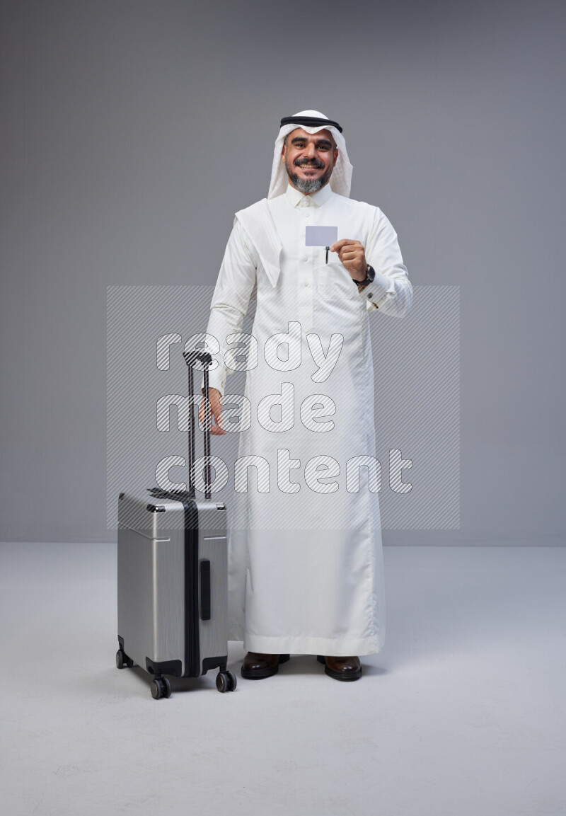 Saudi man wearing Thob and white Shomag standing holding Travel bag and ATM card on Gray background