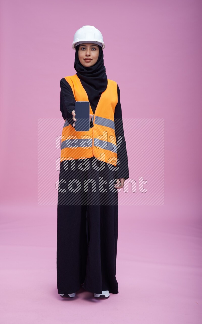 Saudi woman wearing Abaya with engineer vest and helmet standing showing phone to camera on pink background