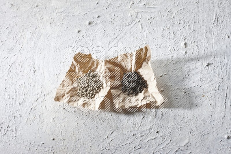 2 Crumpled pieces of paper full of black and white pepper beads on a textured white flooring