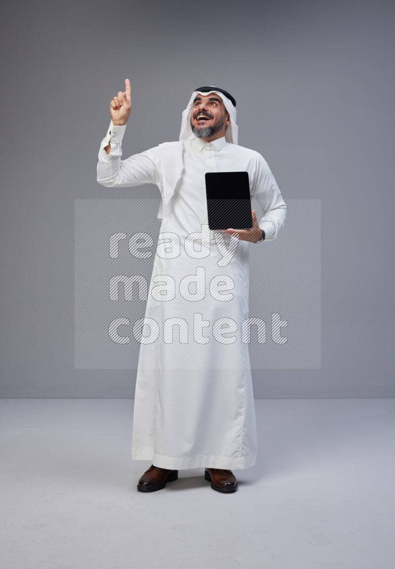Saudi man Wearing Thob and white Shomag standing showing tablet to camera on Gray background