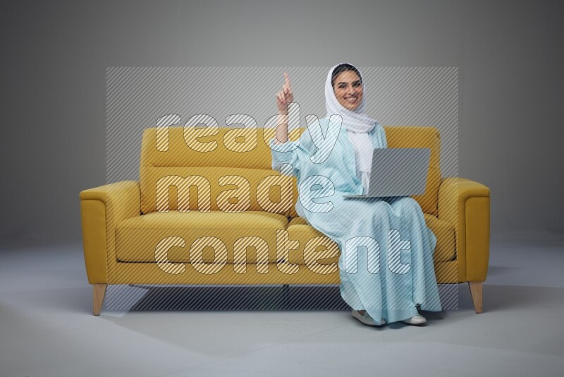 A Saudi woman sitting on a yellow Sofa With her Laptop Pointing wearing Blue Abaya