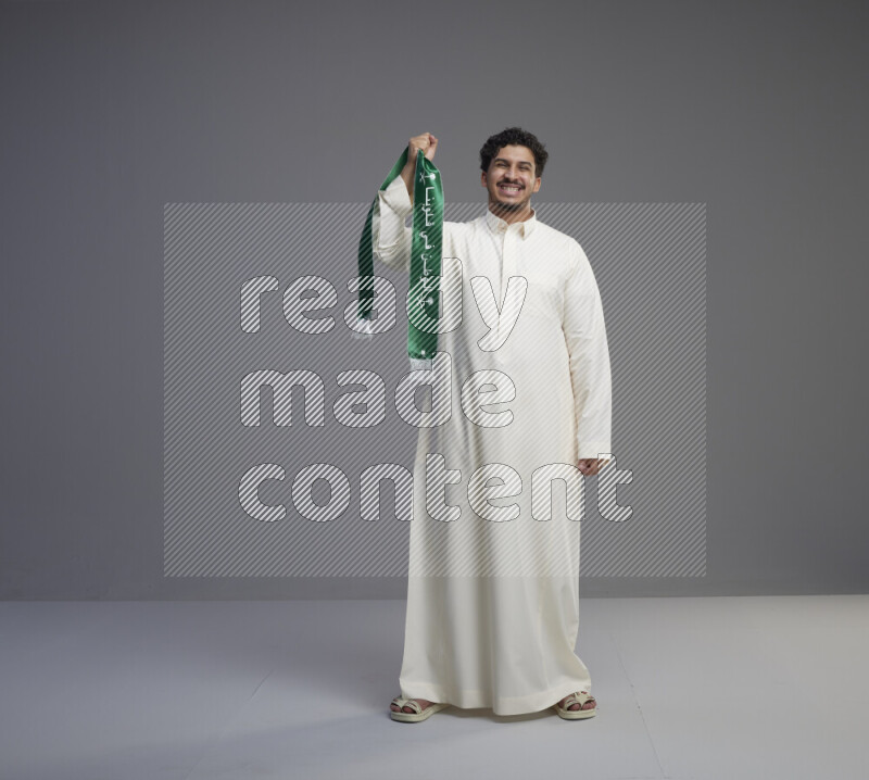 A Saudi man standing wearing thob holding Saudi flag scarf on gray background