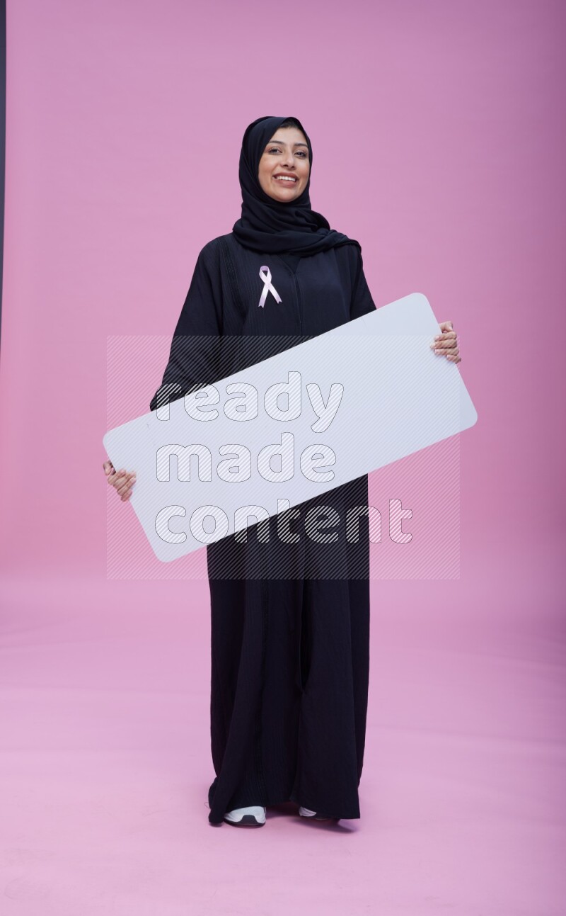 Saudi woman wearing pink ribbon on Abaya standing holding board on pink background