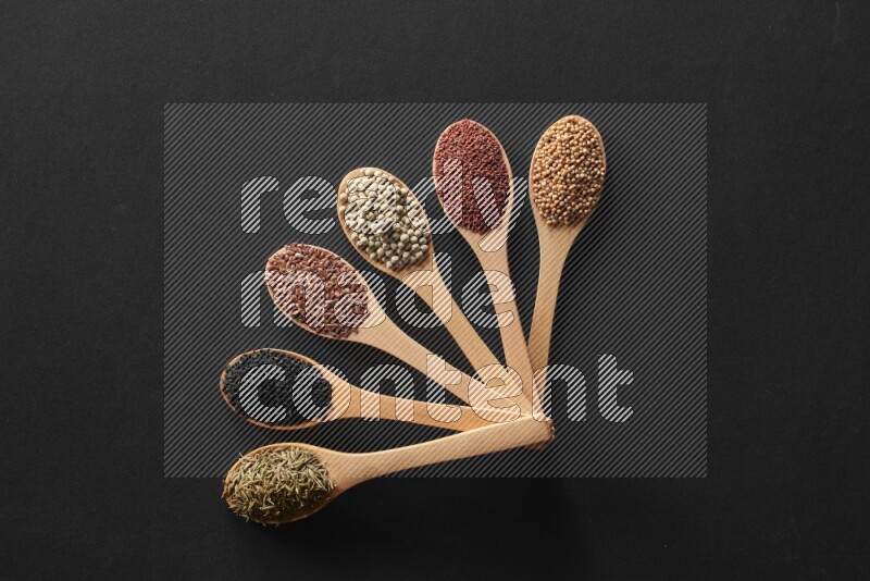 wooden spoons filled with white peppers, mustard seeds, black seeds, garden cress, cumin and flax on black flooring