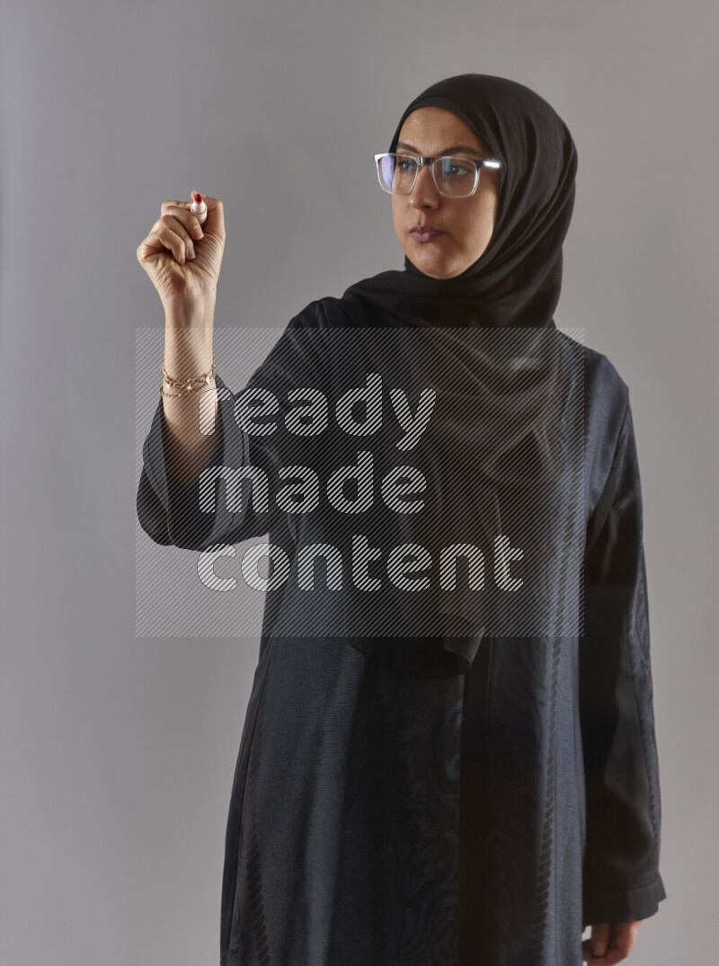 Woman in black abaya, hijab and glasses holding a marker pen to write on a transparent board with different reactions and expressions on grey background