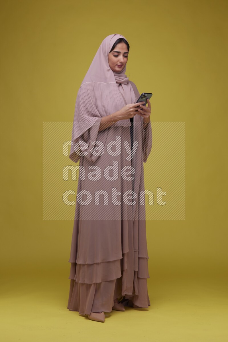 A woman Texting on her phone on a Yellow Background wearing Brown Abaya with Hijab