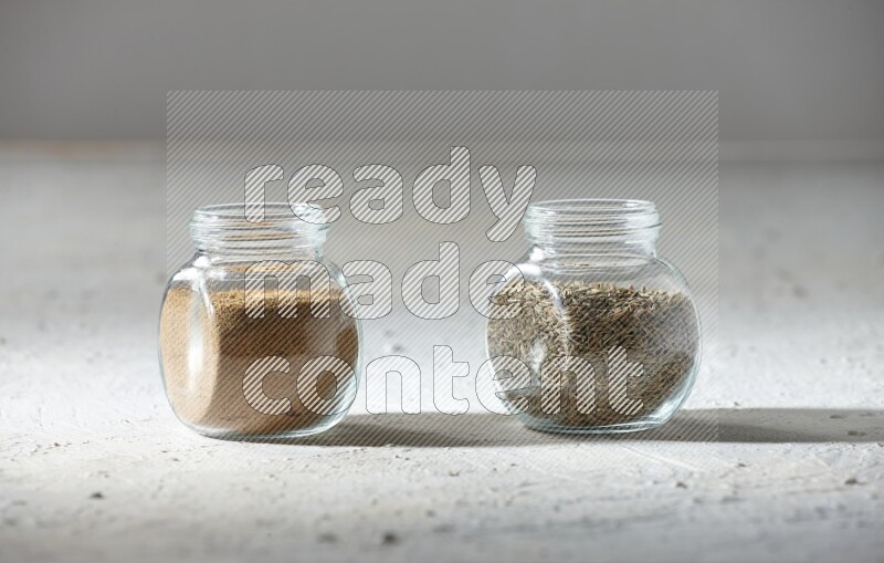 2 glass spice jars full of cumin powder and cumin seeds on textured white flooring