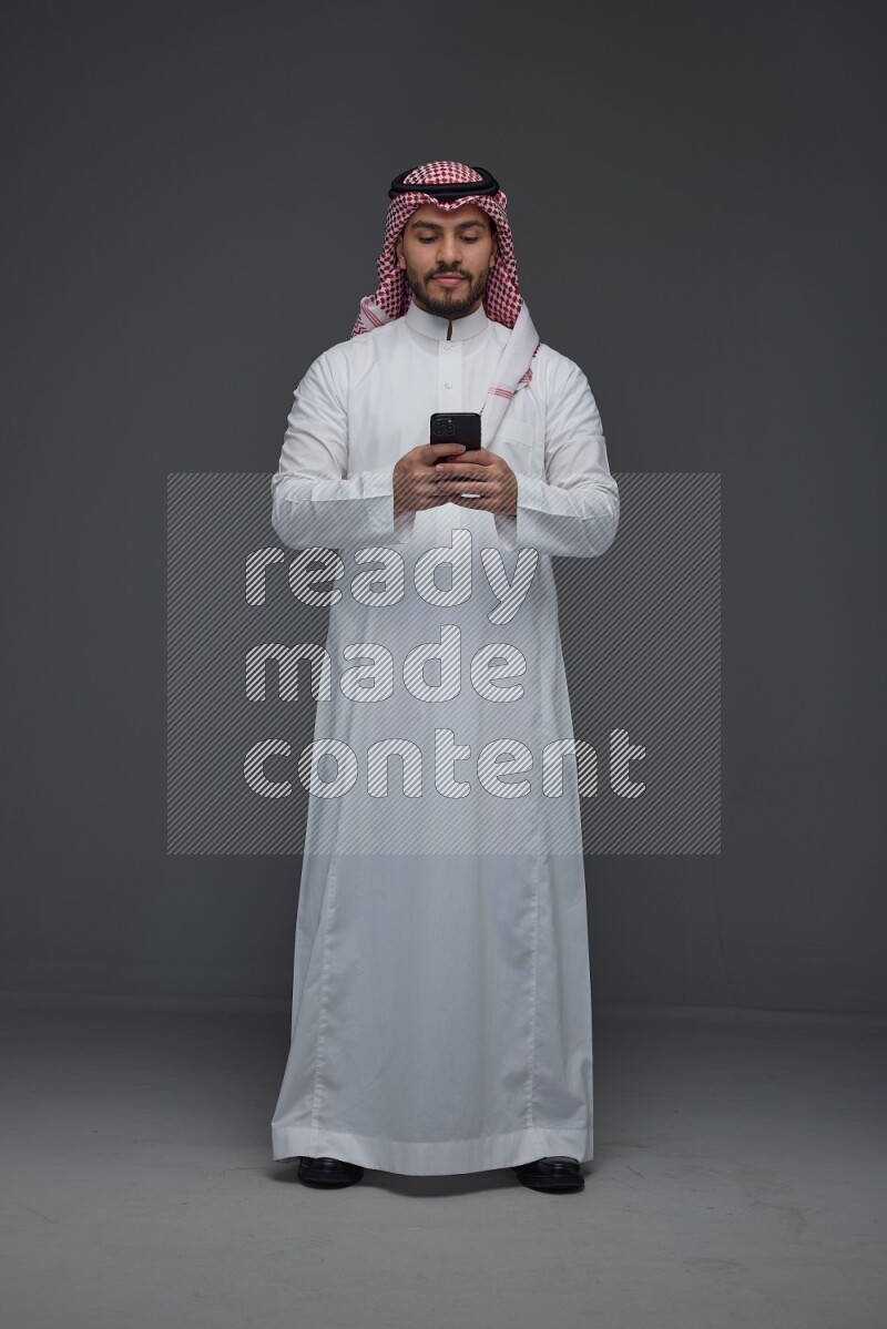 A Saudi man wearing Thobe and Shmagh standing and using his phone eye level on a gray background