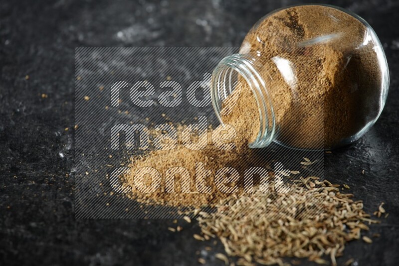 A flipped glass spice jar full of cumin powder with spilled powder and cumin seeds on a textured black flooring