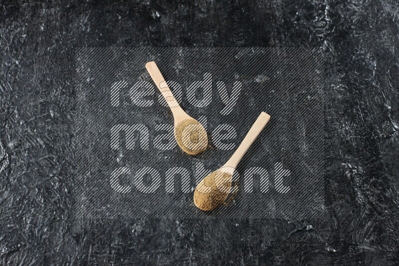 2 wooden spoons full of cumin powder on textured black flooring