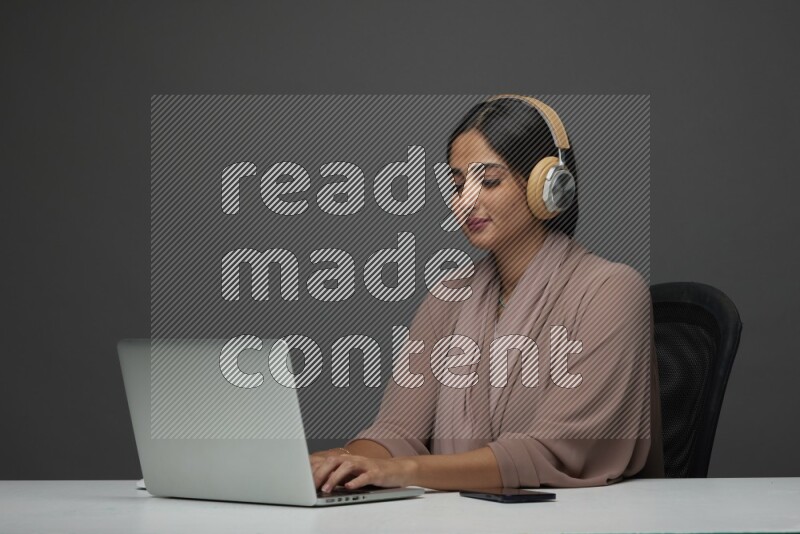 A Saudi woman Sitting on her desk Typing on her laptop wearing a headset  on a Gray Background wearing Brown Abaya