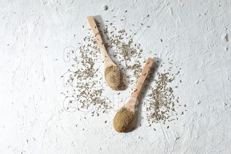 2 Wooden spoons full of cumin powder and cumin seeds on textured white flooring