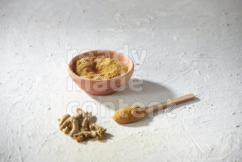 A wooden bowl and wooden spoon full of turmeric powder with dried turmeric fingers on textured white flooring