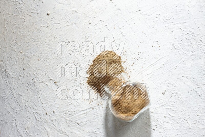 A flipped glass spice jar full of cumin powder and powder spilled out on textured white flooring