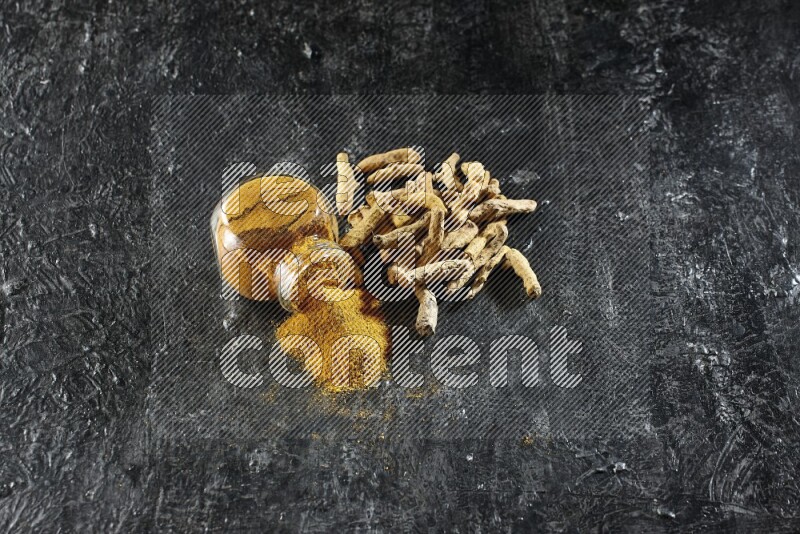 A flipped glass spice jar full of turmeric powder and powder spilled out of it with dried whole fingers on textured black flooring