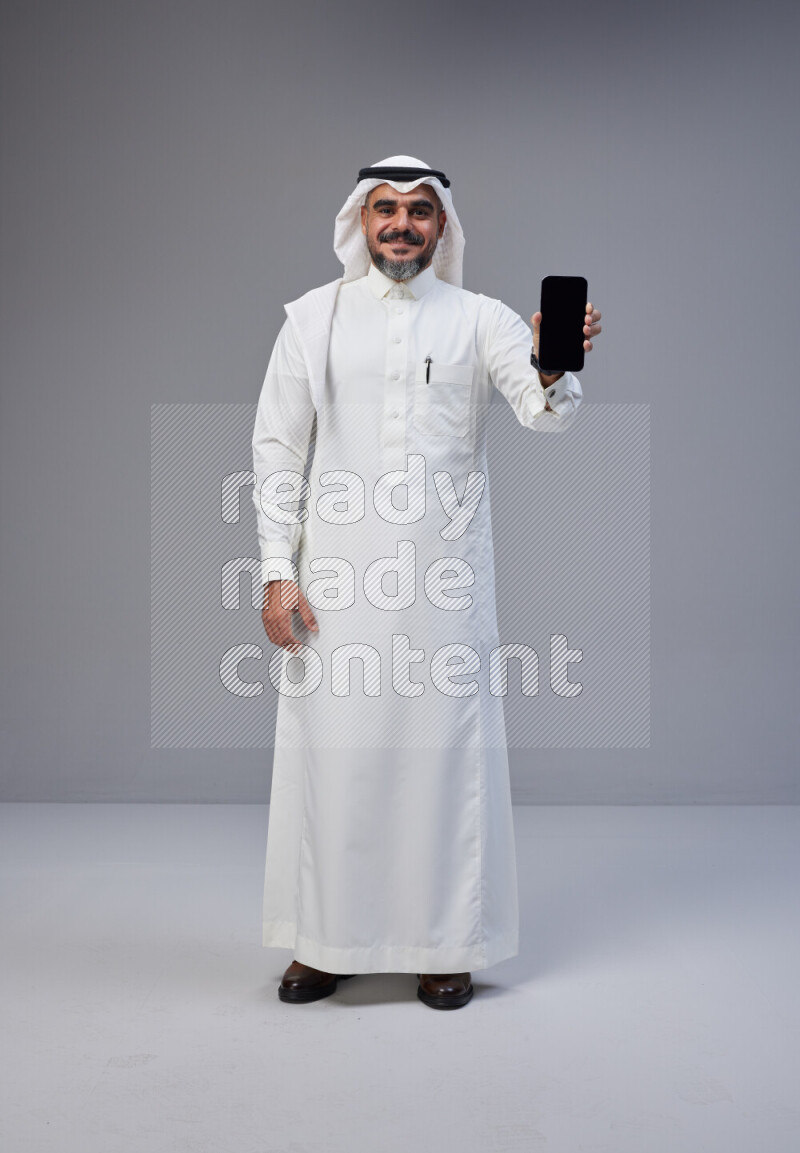 Saudi man Wearing Thob and white Shomag standing showing phone to camera on Gray background