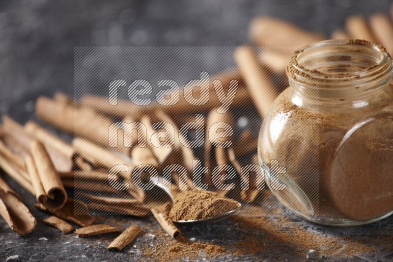 Herbal glass jar and a metal spoon full of cinnamon powder surrounded by cinnamon sticks on textured black background