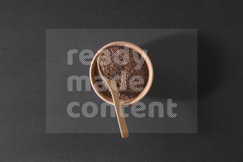 A wooden bowl full of flaxseeds with wooden spoon full of the seeds on it on a black flooring